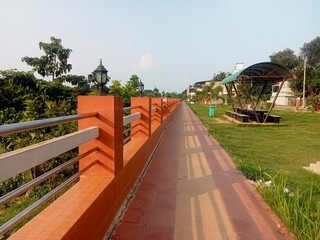 Image depicts an outdoor scene featuring a long, straight pathway with a red and beige tiled surface, bordered by a vibrant orange brick wall with metal railings. The wall is interspersed with black, 