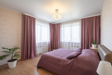 cozy bedroom with two windows draped in mauve curtains and lace sheers.  A bed with matching...