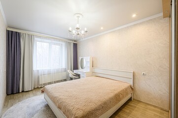 bright bedroom with a bed, dressing table, chandelier, and curtains. The room is neat with calm decor