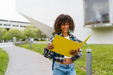 Young hispanic latina college student with afro hair smiling while reading notes from a yellow folder outdoors on campus
