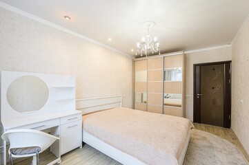 Bright bedroom featuring a bed, wardrobe with mirrors, vanity desk, and a chandelier. Neutral tones...