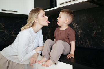 Playful young mother and her little son sticking out their tongues at each other and smiling in a fun and lighthearted moment in the kitchen.