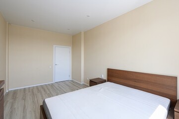 Fototapeta premium Bright bedroom with a wooden bed and white mattress, pale beige walls, and a white door. Minimalist decor