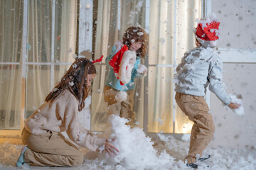 Obraz premium Three kids dressed in festive Christmas clothing enjoy playing with artificial snow on a cozy balcony, capturing the warmth and fun of the holiday season at home in a joyful moment.