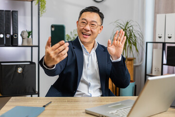 Happy excited Asian middle-aged businessman blogger influencer making phone conversation call with social media followers at office workplace working on laptop. Freelancer Chinese man in formal suit