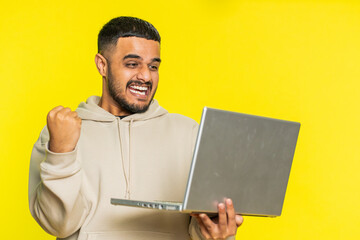 Excited happy Indian man typing on laptop computer, working on project, enjoying results, winning lottery game, celebrating success, online shopping, victory. Arabian guy on yellow background indoors