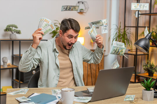 Rich Caucasian man earnings big profit success win lottery money cash rain falling shopping more tips. Happy young freelancer guy manager with laptop clenching fists at home office desk. Lifestyle.