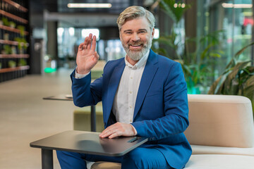 Hello. Happy Caucasian businessman employee waves hand hi gesture greeting welcomes someone, online webinar, remote distance conversation. Entrepreneur man guy in suit sitting in modern office lobby. © Andrii Iemelianenko