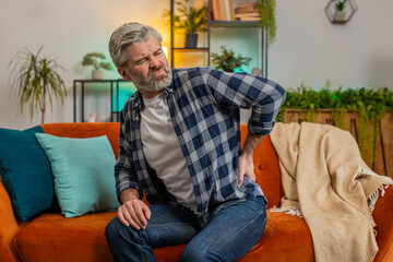 Fototapeta premium Tired senior bearded man suffering from back pain sitting on sofa in living room at home. Sad old male massages painful lower back muscles. Caucasian mature guy exhausted overworked in apartment.