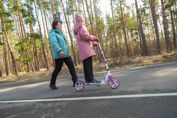 Little girl riding scooter while walking with her grandmother in the forest on a sunny day