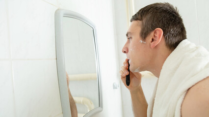 Fototapeta premium Young man with naked torso shaving his beard with foam looking at his reflection in mirror at bathroom
