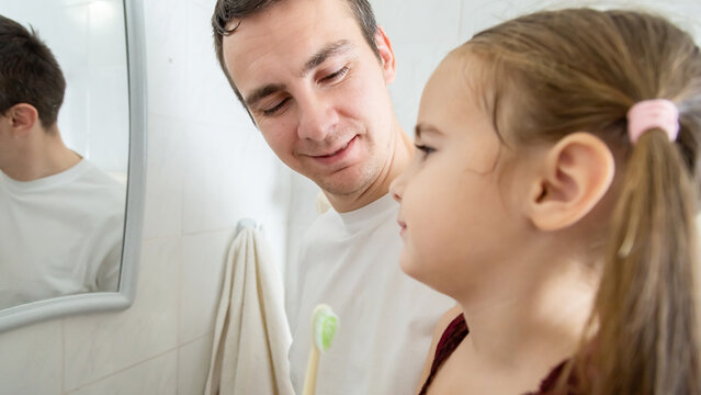 A loving father is holding his daughter in the arms and helping her brushing their teeth in a bathroom. - Powered by Adobe