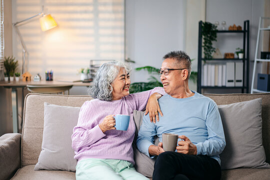 Senior Asian Couple Relaxing on Sofa at Home Enjoying Coffee or Tea Smiling Together Happy Retirement Lifestyle Concept Spending Time Together