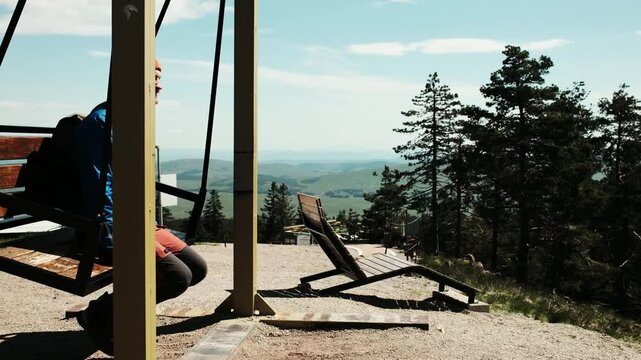 A man with a backpack sits on a wooden swing with mountain views at Tornik, Zlatibor. Spring day, peaceful atmosphere. Solo travel concept. Horizontal 4k footage