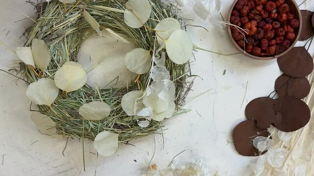 A woman thoughtfully arranges dried flowers on a handmade wreath base, planning the floral composition before attaching them. Her hands carefully place elements like lavender, statice, wheat, eucalypt