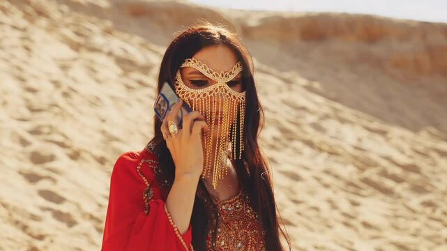 glamour arabian woman hands holding smartphone. red gold oriental abaya dress black long hair sand desert. sexy girl arab style beauty face hides golden mask burka talking on the phone. summer nature