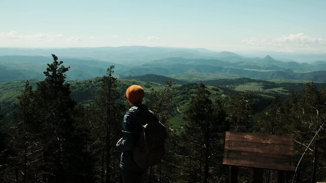 Young woman hiker with backpack and orange hat stretches her arms wide while enjoying a panoramic mountain view from Tornik peak in Zlatibor, Serbia during spring. Solo travel concept
