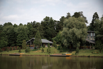 Obraz premium Peaceful lakeside scene with charming cottages nestled among lush greenery. The tranquil setting features a calm lake, dense forest, and inviting docks perfect for relaxation.
