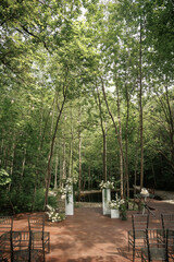 Obraz premium A picturesque outdoor wedding venue set in a forest surrounded by tall trees. The scene includes elegantly arranged chairs and floral decorations, creating a serene and romantic atmosphere.