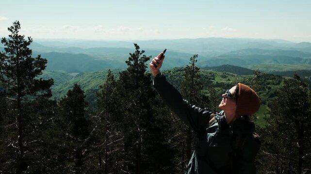 Female traveler taking selfie with breathtaking spring landscape in the background, standing confidently above the forest in Zlatibor, Serbia country. Travel blogger making video outdoor