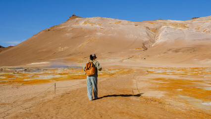 A lone traveler explores the vibrant geothermal area in the Icelandic highlands. The landscape is characterized by colorful mineral deposits and rugged terrain, under a bright blue sky.