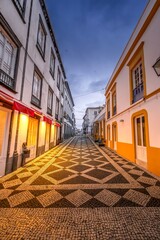 A beautiful cobblestone street with a distinctive mosaic pattern stretches through Ponta Delgada, Azores, illuminated by warm lights at dusk. The traditional buildings showcase Azorean architecture