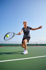 A senior woman is swinging her racket while hitting a tennis ball during a bright, sunny afternoon at an outdoor tennis court. Healthcare and active retirement concept. Generative AI.