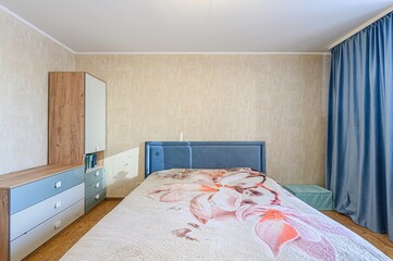 cozy bedroom features a bed with a floral quilt, a blue headboard, a cabinet, and blue curtains....