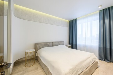 Bright bedroom featuring a gray bed with white linens, gold side tables, blue curtains, and warm...