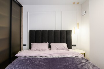 modern bedroom featuring a dark upholstered headboard, soft bedding, gray curtains, and a unique circular light fixture. The room has white walls with paneling and a light wood floor