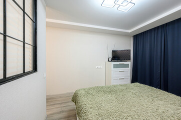 bedroom features a bed with a green fuzzy blanket, a window, a TV on a white dresser, and blue curtains