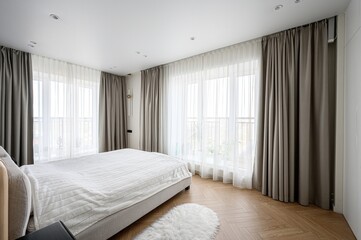 bright bedroom features a bed with a striped white cover, flanked by grey curtains and sheer white...