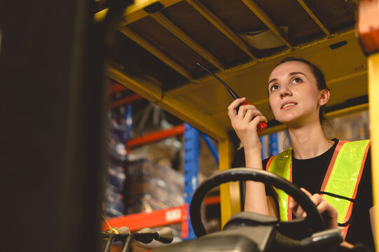 warehouse, radio, and forklift in shipping warehouse. Female transport worker in safety uniform transport worker talking on walkie talkie, drives forklift at freight cargo warehouse port.