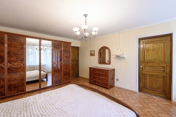 bedroom featuring a large bed with a white cover, a wooden wardrobe with mirrored doors, a dresser with a mirror above, and a chandelier. The room has wooden floors