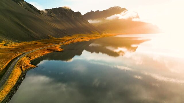 Vulkanische K&uuml;stenlandschaft in Island im goldenen Morgenlicht mit ruhiger Wasserfl&auml;che, Spiegelung der Berge und einer schmalen Stra&szlig;e entlang des Ufers in herbstlicher Umgebung