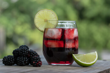 A glass of blackberry juice with ice and lime garnish on a wooden surface with blackberries near