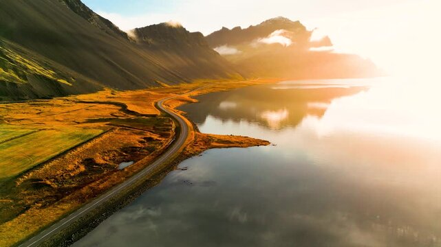 Vulkanische K&uuml;stenlandschaft in Island im goldenen Morgenlicht mit ruhiger Wasserfl&auml;che, Spiegelung der Berge und einer schmalen Stra&szlig;e entlang des Ufers in herbstlicher Umgebung