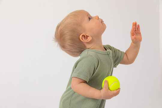Side view one year little baby boy holding tennis ball and looking up on white background. Sport, childhood and early development concept. Copy space for ad, text. Beach summer tennis camp