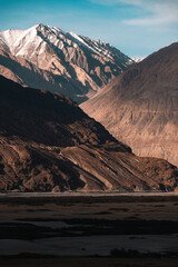 Golden sunlight casts long shadows across the dramatic Nubra Valley landscape, revealing rugged mountain textures and snow-capped Himalayan peaks under a vast blue sky in northern Ladakh, India