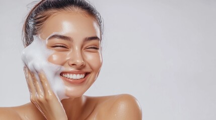 In a skin care advertisement, a woman smiles while washing her face with white foam on a white background. The skin texture is shiny and healthy, and the skin tone is naturally glowing. 