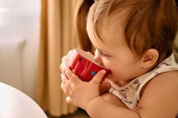 A close-up of a baby with bright blue eyes, wearing a white patterned onesie, drinking from a red...