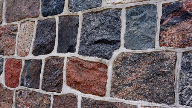 Close-up view of a multicolored stone wall with irregular rectangular blocks and white grout. The textured surface and natural colors give a rustic and architectural feel.