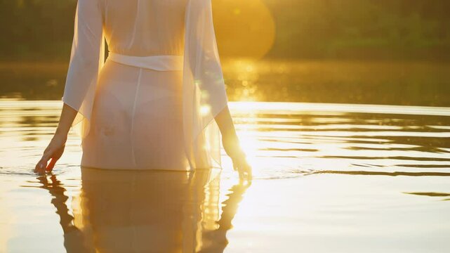 happy fantasy woman river nymp enjoys freedom magic warm sun light hot summer green nature. Girl elf stands in water hands strokes lake, wet white long silk dress back rear view. Sunset yellow flare