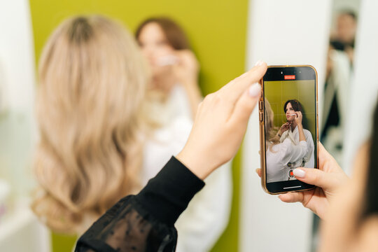 Close-up of female hands holding smartphone recording video of beautician applying makeup on model face, creating content for social media or online tutorial. Concept of beauty treatment.