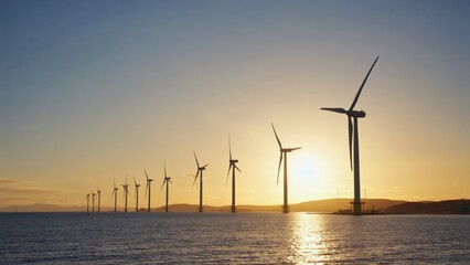 Obraz premium Offshore wind turbines stretch across the calm sea at sunset, their blades turning slowly against the golden sky, symbolizing clean energy, sustainability and the future of renewable power generation.