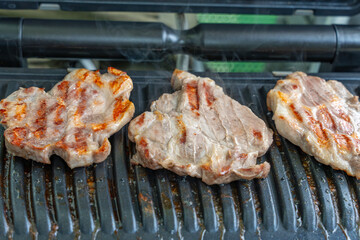 Three juicy pieces of meat are grilling on a contact grill, showing distinct grill marks. Light smoke rises as the meat sizzles.