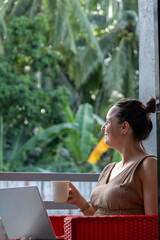 A woman in a natural dress sips her morning coffee on a hotel balcony, absorbed in her laptop,...