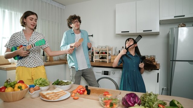 Caucasian skilled father, mother and asian daughter making breakfast while dancing together. Skilled mom playing ukulele while preparing vegetable at modern kitchen. Healthy food concept. Pedagogy.