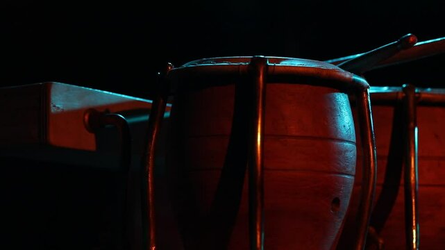 A cinematic close-up of Latin timbale drums under dramatic red and blue stage lighting. The camera tilts up and pulls focus to an atmospheric, blurry shot.