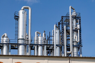 distillery installation with multiple distillation columns and pipelines. The metal structure rises above the factory roof, set against a clear blue sky.